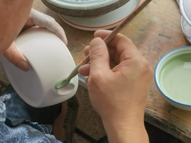 A craftsman hand-painting sage green glaze onto the handle of a white ceramic mug with a fine brush, with a bowl of green glaze beside them on a wooden workbench