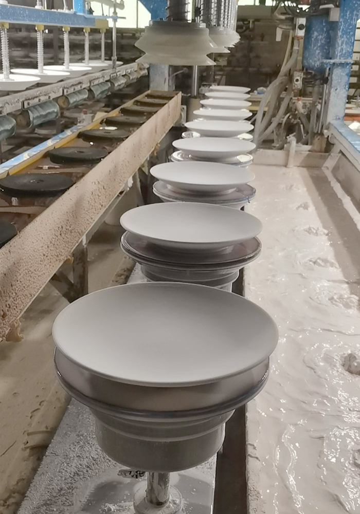 A row of white ceramic plates mounted on rotating spindles along an automated glazing conveyor line inside a porcelain manufacturing factory